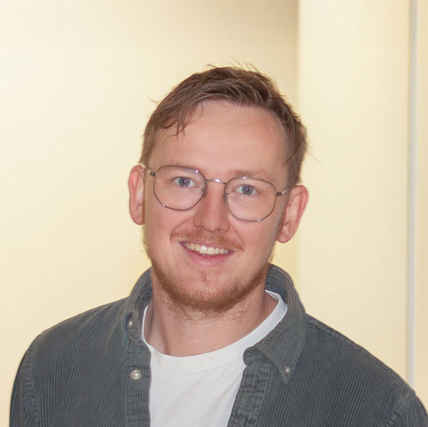 Porträt von Lars Rußwurm, Mitarbeiter der Stiftung Heilpädagogisches Kinderhaus gGmbH. Er trägt eine runde Brille, ein helles T-Shirt und ein offenes graugrünes Hemd. Er lächelt freundlich in die Kamera. Der Hintergrund ist hell und ruhig, die Atmosphäre wirkt offen, zugewandt und professionell.